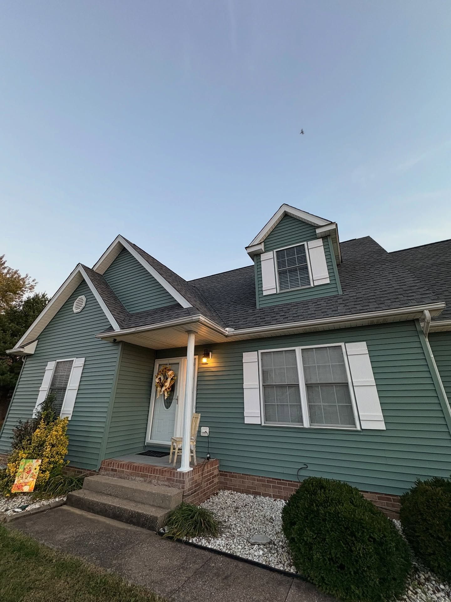 Green-sided suburban house with white trim, front porch, dormer windows, and manicured shrubs at dusk