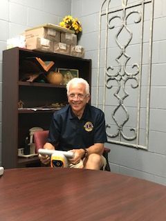 Man seated at a desk, smiling, wearing a Lions Club shirt. He holds a device. A shelf with items is behind him.