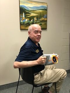 Man with a bald head holds an orange device, sitting in a chair, smiling at the camera. Painting on the wall behind him.