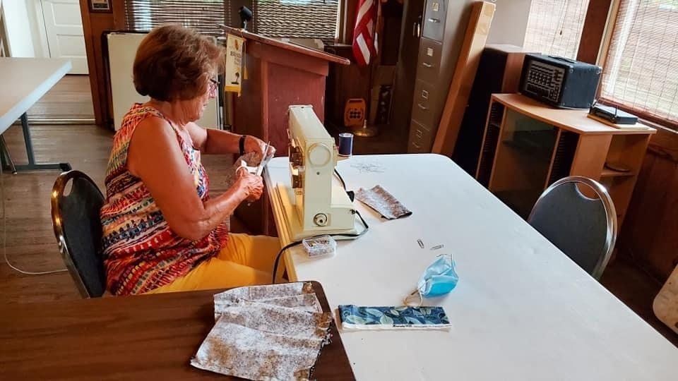 Woman sewing at a table in a room, with a sewing machine, fabric, and other materials.