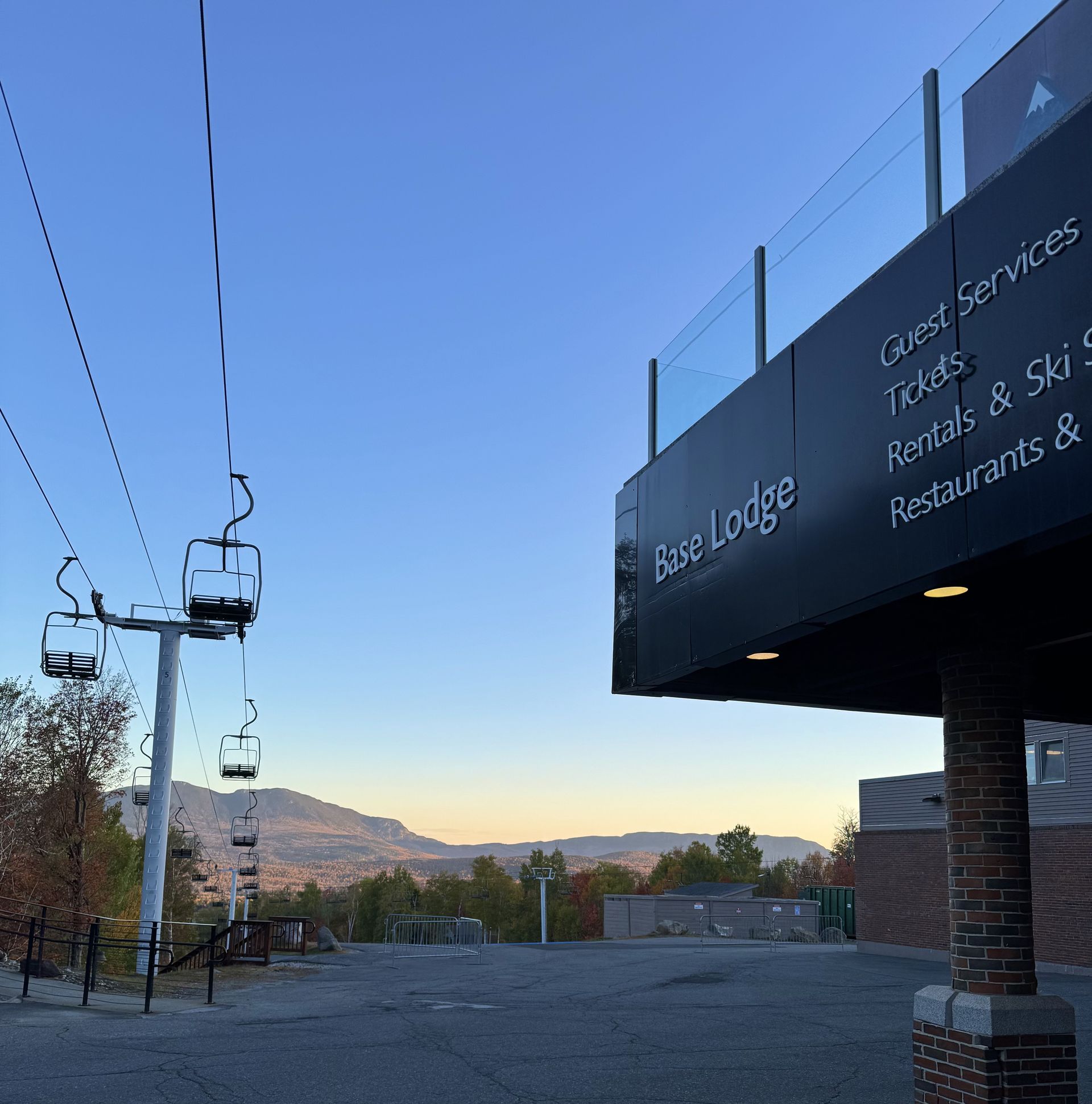 Ski lodge sign beside a gondola lift at dusk, with mountains and a blue sky in the background