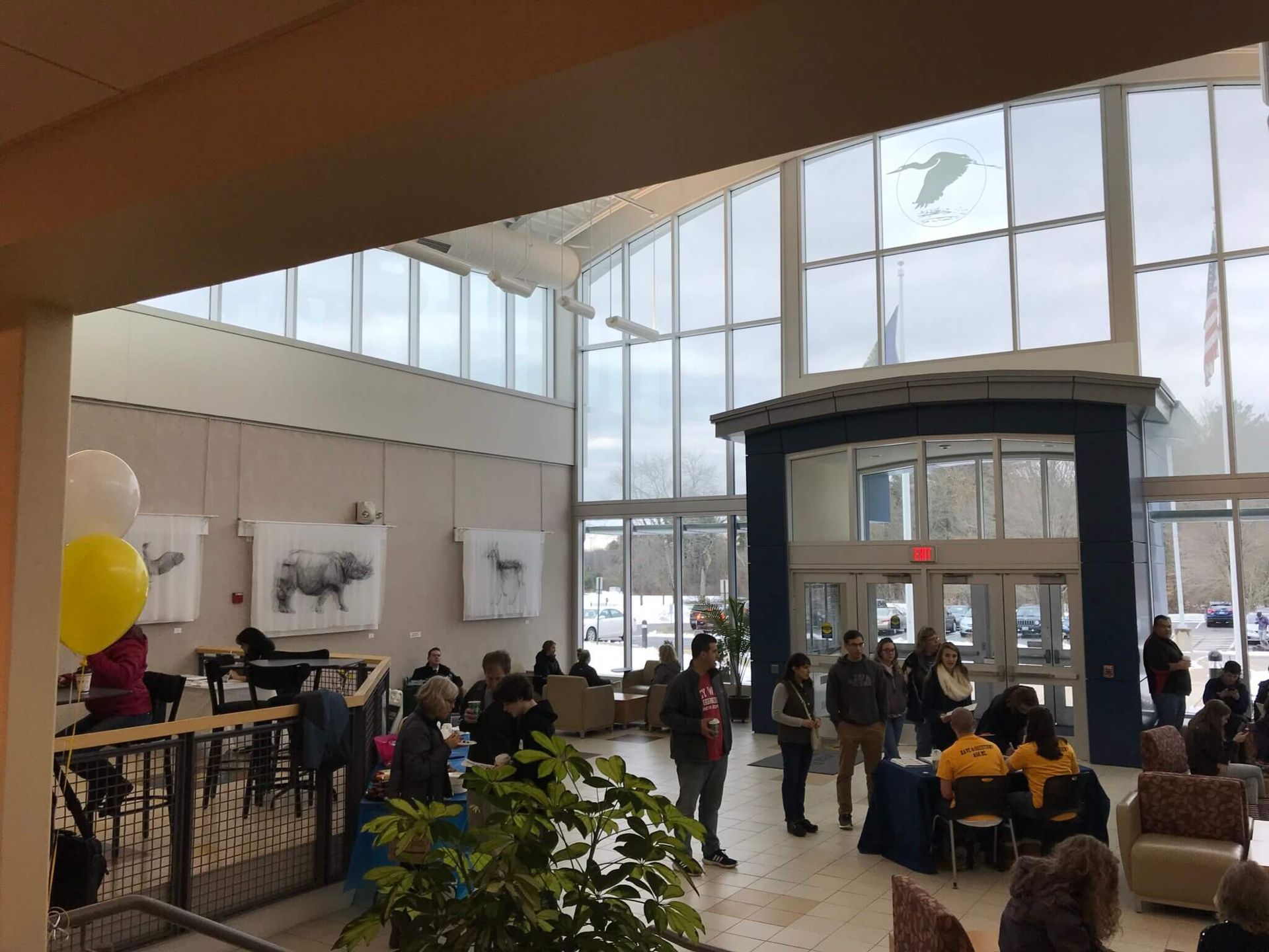 People gather in a bright, high-ceilinged lobby with tall glass windows, indoor plants, and artwork on the walls.