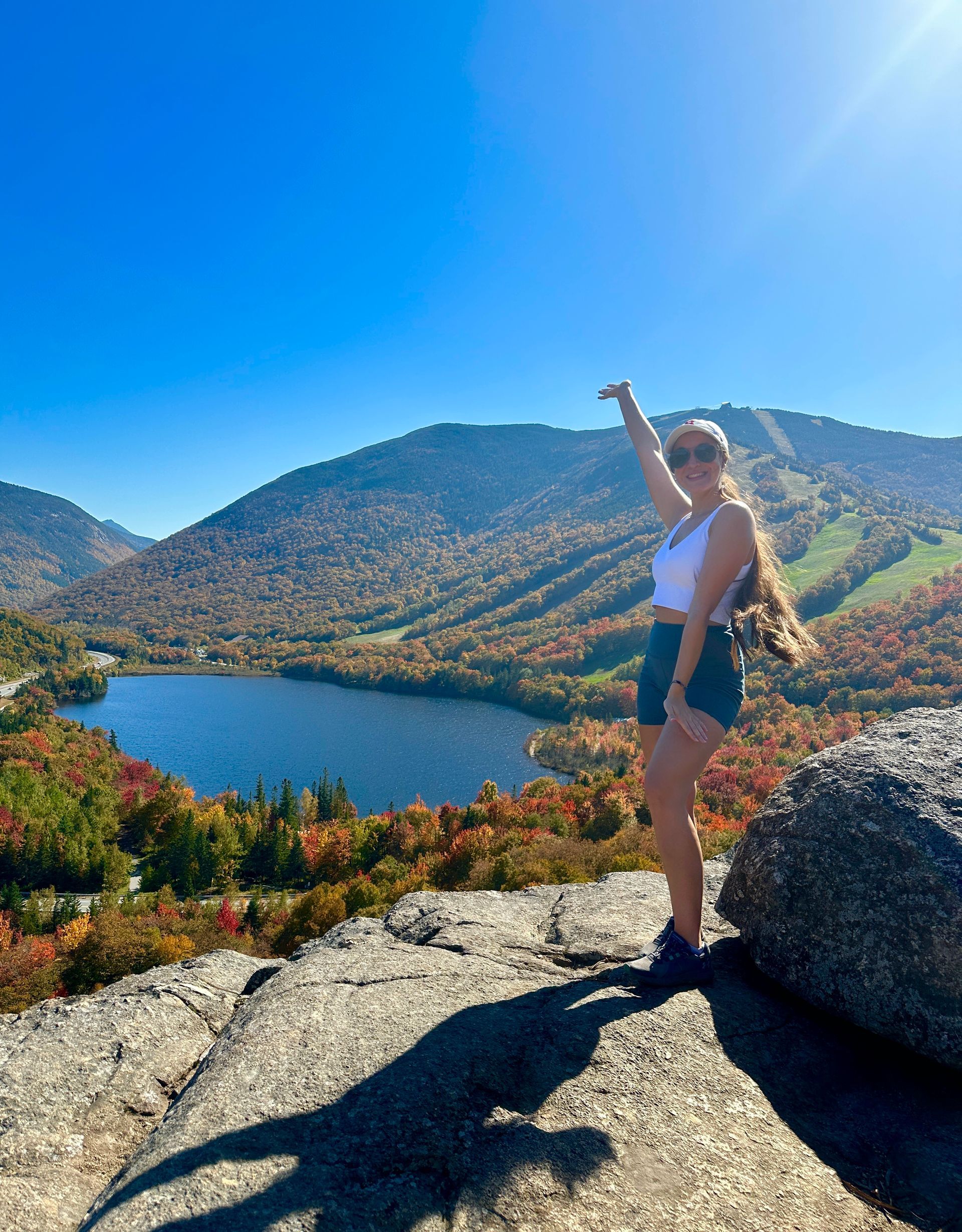 landscape artist Nicole Ann standing on a ledge of a mountain in New Hampshire that showcases a colorful mountain scene with a lake and fall foliage after a hike up Artists Bluff hiking trail