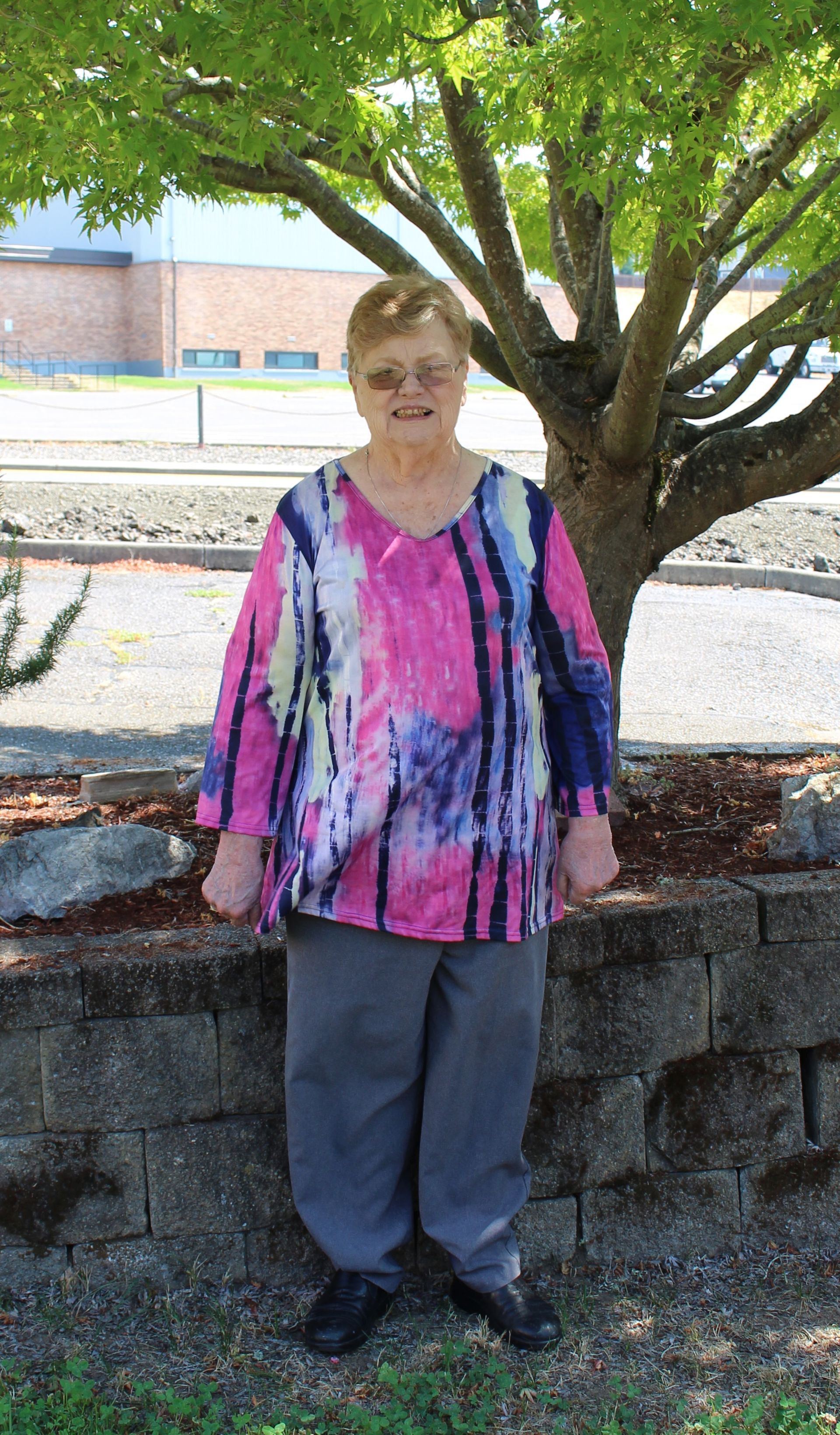 Woman in tie-dye top and gray pants stands in front of a tree and brick wall.