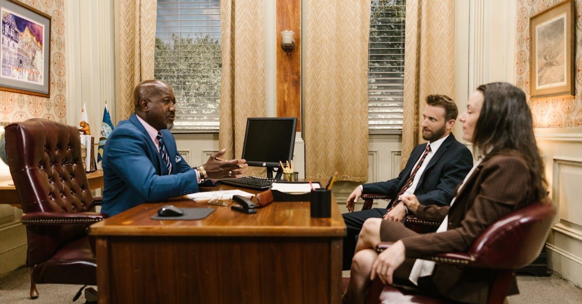 Three people seated across a desk in a formal office, talking during a meeting.