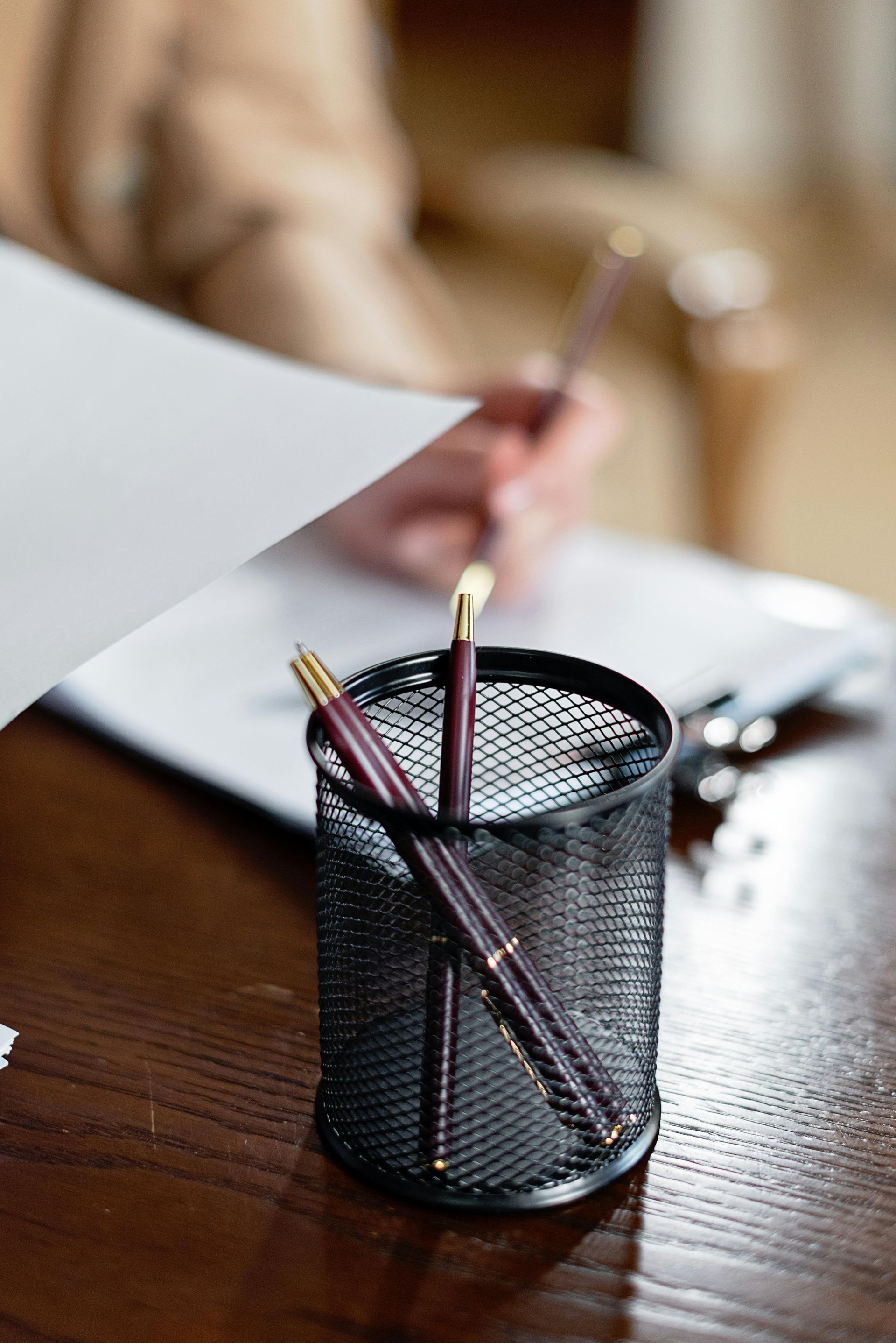 Black mesh pen holder with pens and a pencil on a wooden desk, with a blurred person writing in the background