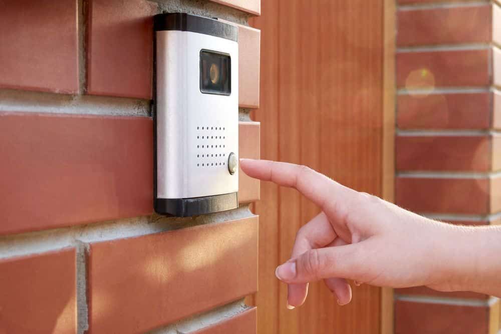 Hand Pressing a Doorbell on a Brick Wall Beside a Wooden Door — IRIS CCTV in Grafton, NSW