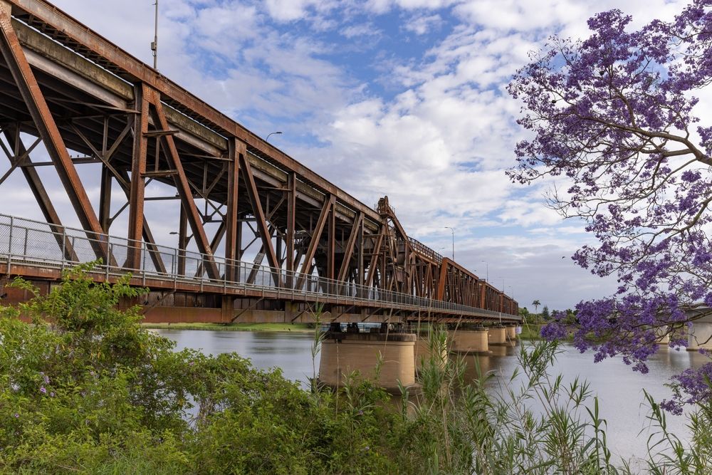 Rust-colored Metal Bridge in Grafton Spanning a River, Flanked by Greenery and a Tree With Purple Blooms — IRIS CCTV in Grafton, NSW