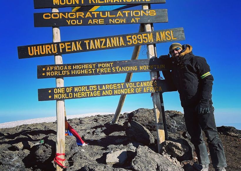 Climber posing by Mount Kilimanjaro summit sign on rocky peak under bright blue sky