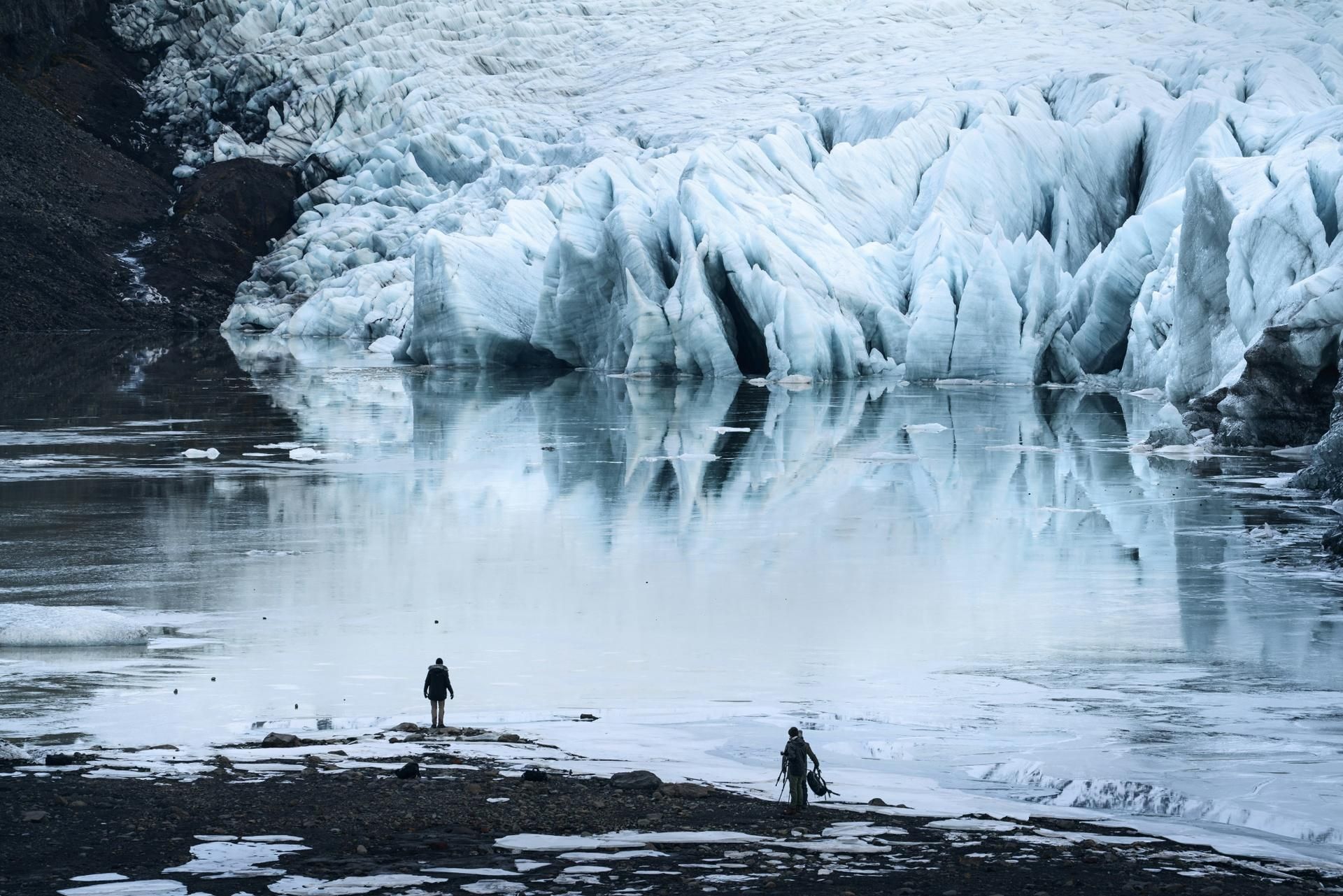 Two people on a rocky shore before a blue glacier reflecting in still water