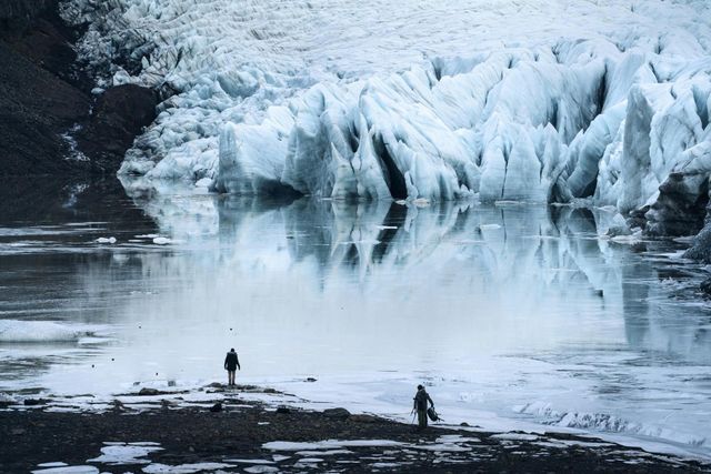 Two people stand on a rocky shore before a विशाल blue glacier reflecting in calm water