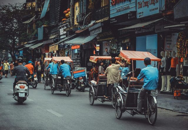 Rickshaws and scooters crowd a busy city street lined with shops and signs.