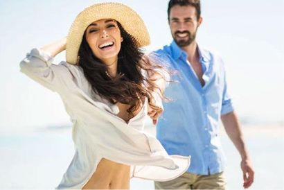 Smiling woman in a sunhat walking on a beach with a man in a blue shirt behind her