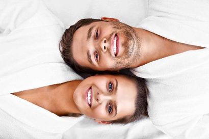 Two smiling people lying on white pillows, heads together, looking at the camera.