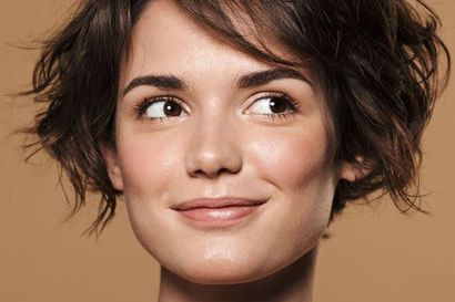 Close-up portrait of a smiling person with short wavy brown hair against a beige background