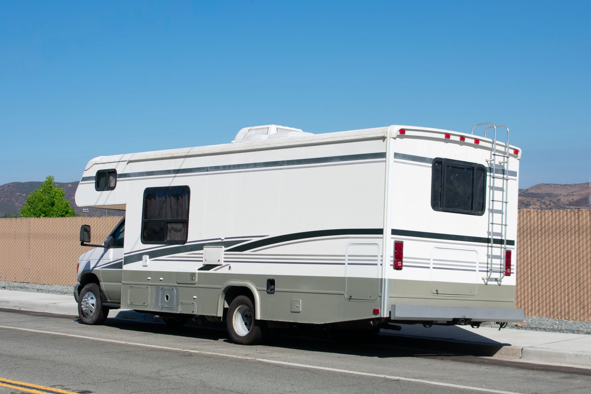 White and tan RV parked on a road beside a tan wall, under a clear blue sky.