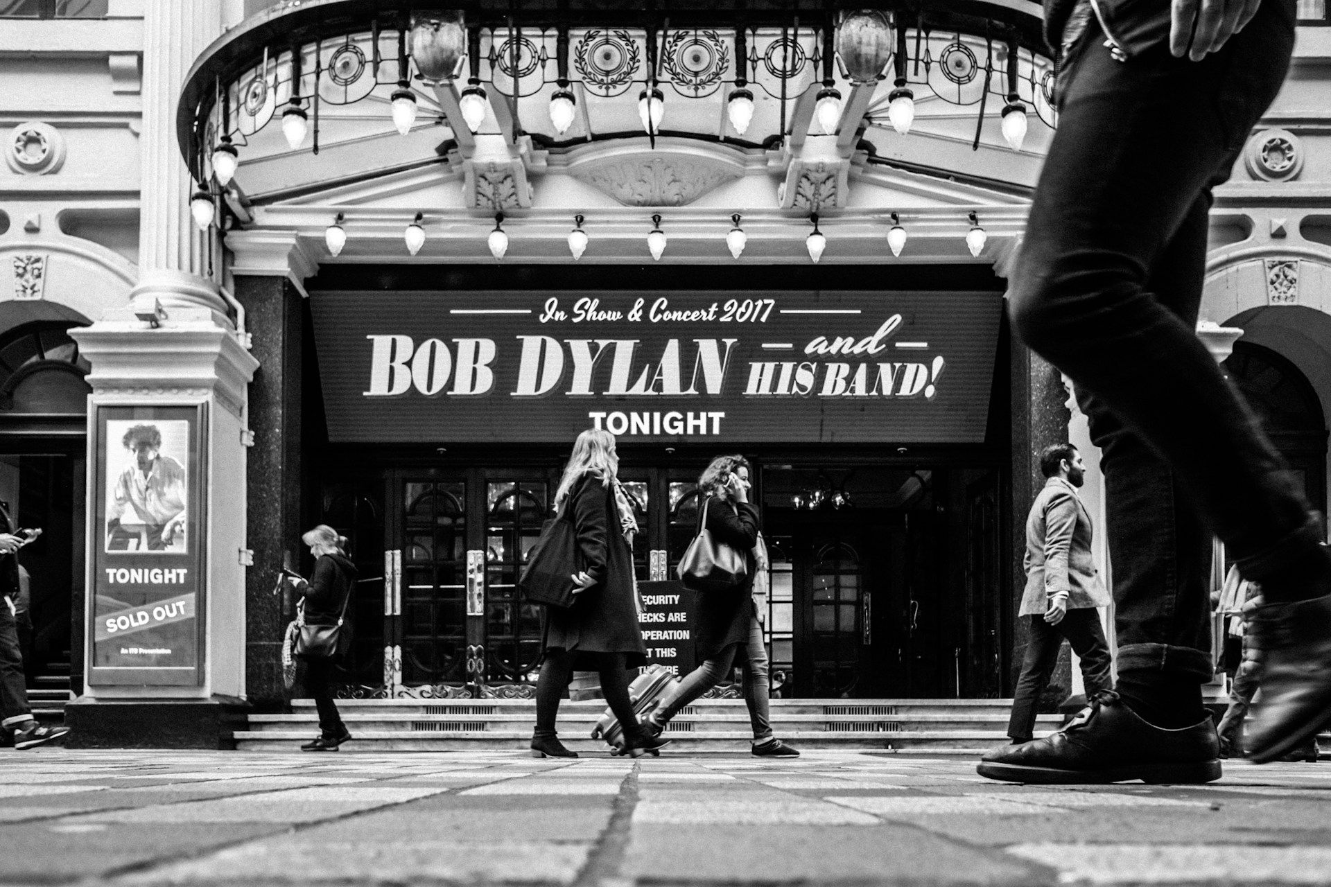 A black and white photo of people walking in front of a bob dylan concert.