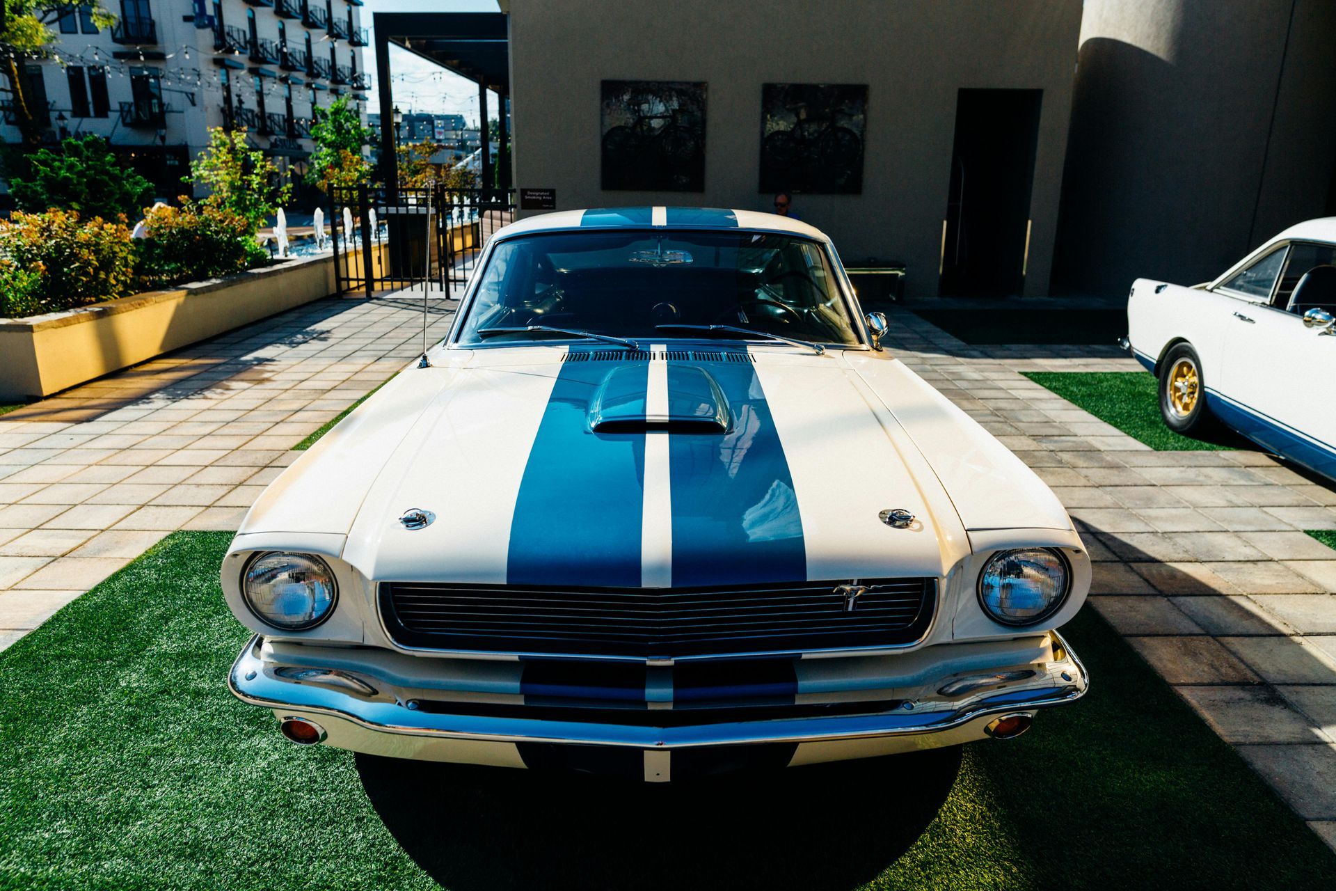 A white mustang with blue stripes on the hood is parked in front of a building.