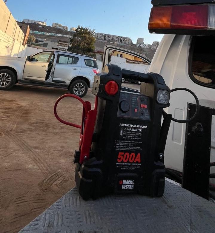 Una persona agachada al borde de la carretera conecta los cables de arranque de una batería portátil a una camioneta verde.