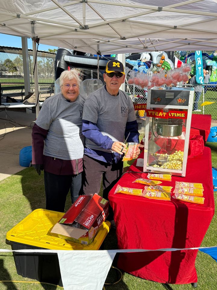 Two people, smiling, behind a popcorn stand at an outdoor event. Red table, machine, and snacks.