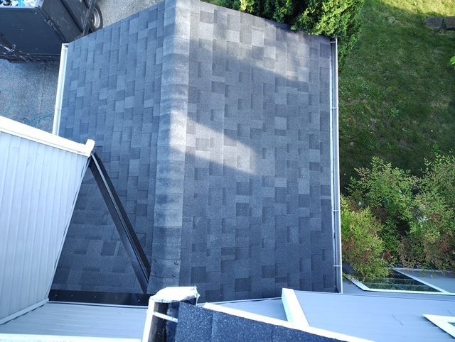 Aerial view of a gray shingled roof with white siding and surrounding green trees.