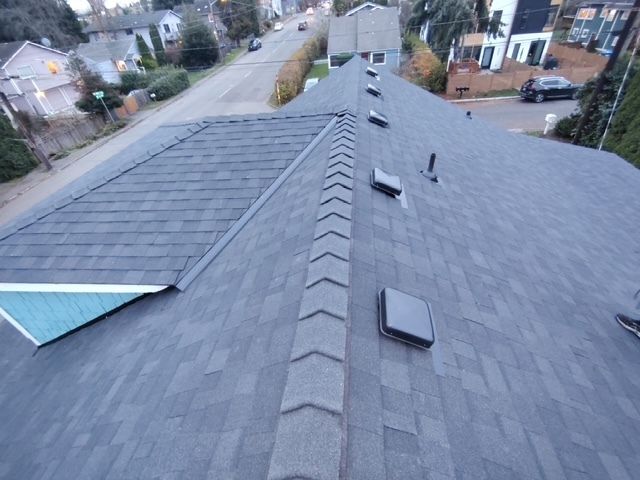 Aerial view of a residential shingle roof with vents and nearby houses in the background