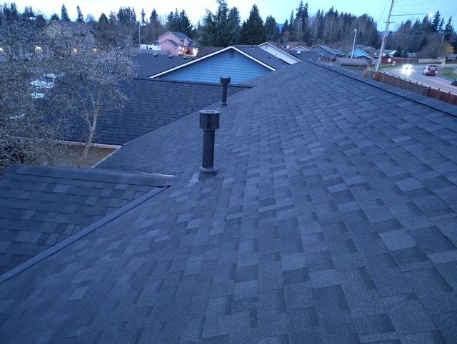 View from a shingled rooftop with a vent pipe, overlooking trees and nearby houses at dusk