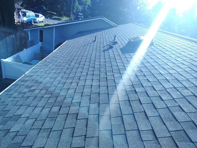 Rooftop with gray shingles and bright sun glare, viewed from above with a house and trees in the background
