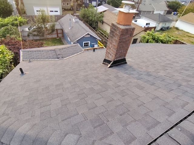 Rooftop view of gray shingles with a brick chimney and nearby houses in the background