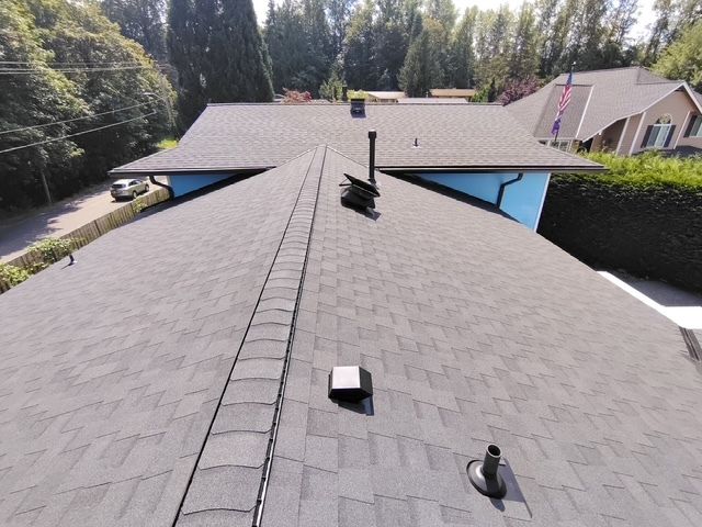 Rooftop view of gray asphalt shingles with vents, chimneys, and neighboring houses in a suburban area