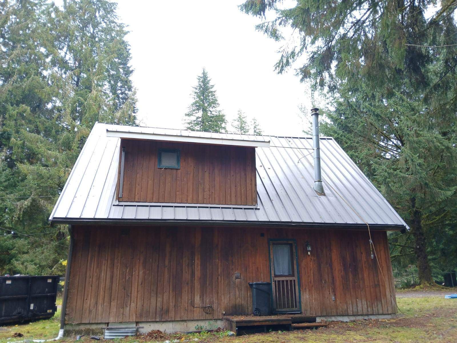 Wooden cabin with metal roof in a forest clearing, with a small porch and chimney.
