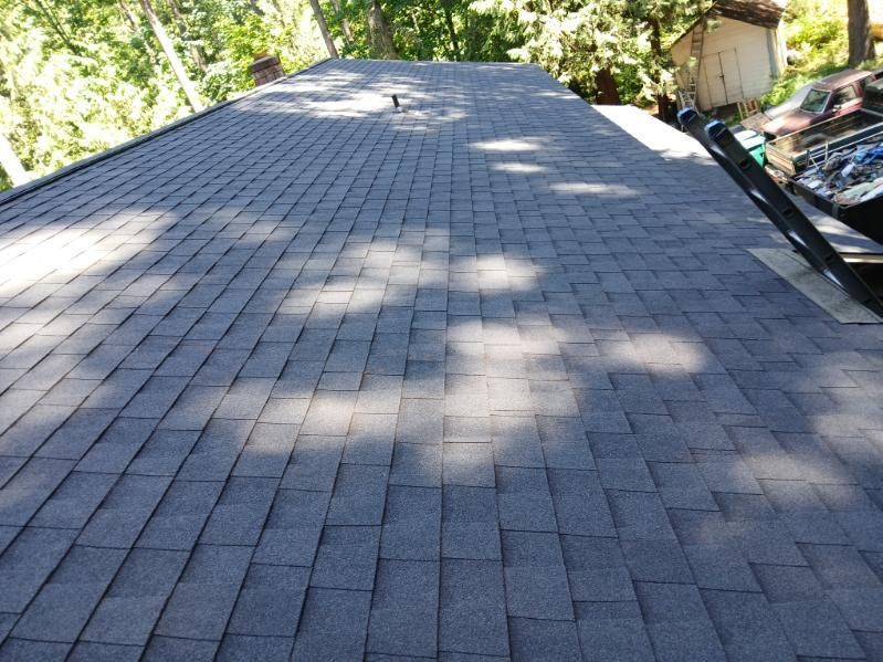 Shingled roof with dark gray asphalt shingles, bordered by trees and a parked car in the background