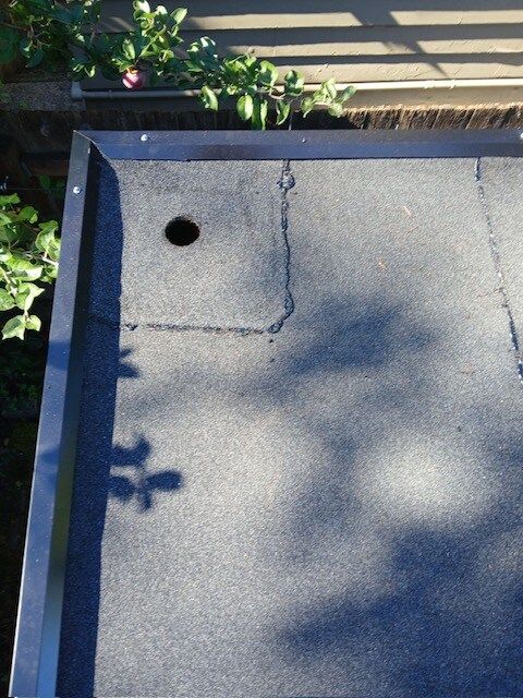 Flat gray roof with a circular vent hole and a visible crack, bordered by plants and wood trim.