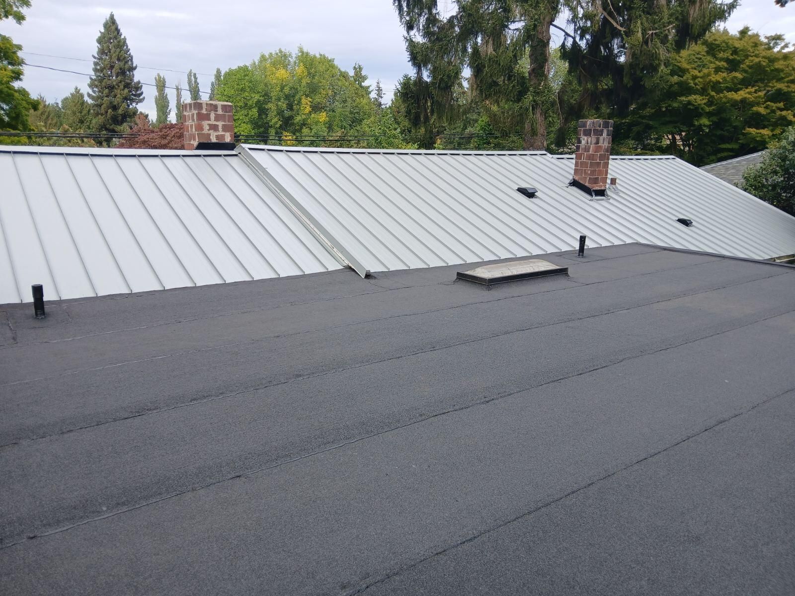 Rooftop with dark shingles in foreground and light metal roof behind, with trees and chimneys in the background