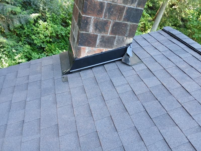 Roof shingles around a brick chimney with a metal flashing strip and surrounding trees visible.