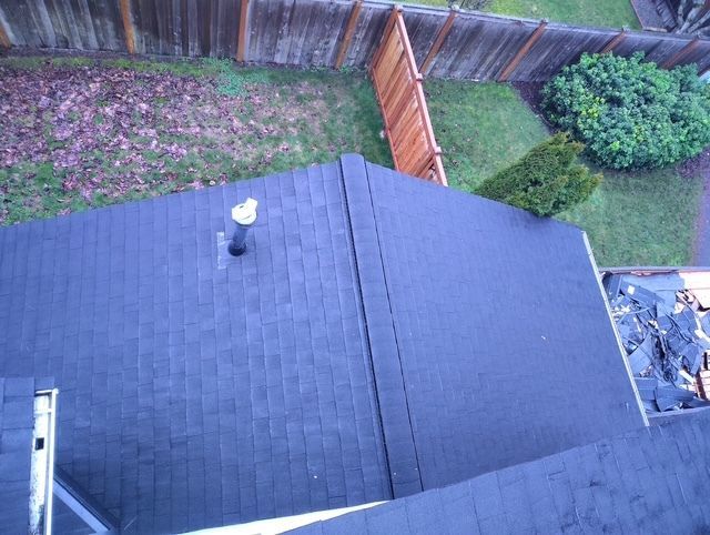 View from above of a dark shingled roof beside a fenced backyard with grass and trees.