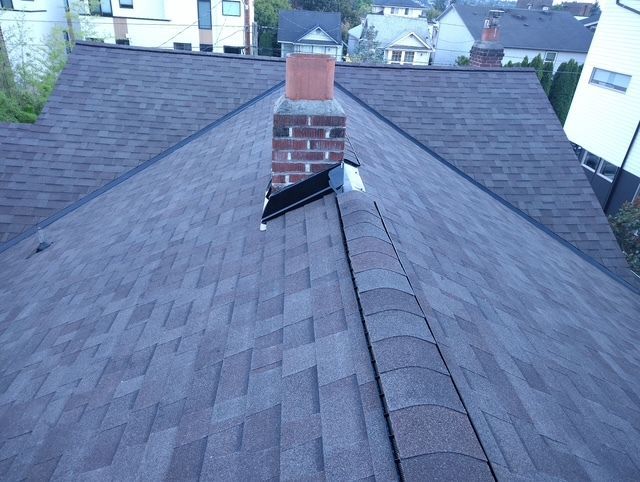 Roof shingles with a brick chimney and a broken black vent or panel near the ridge.