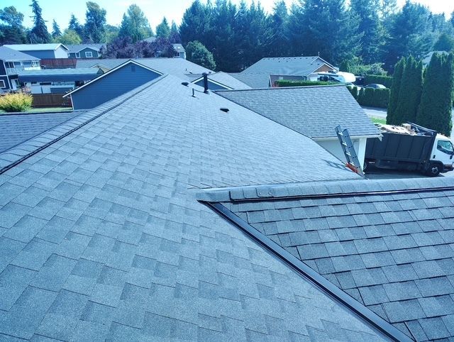 Rooftop under construction with gray shingles and a rooftop unit, trees and houses in the background