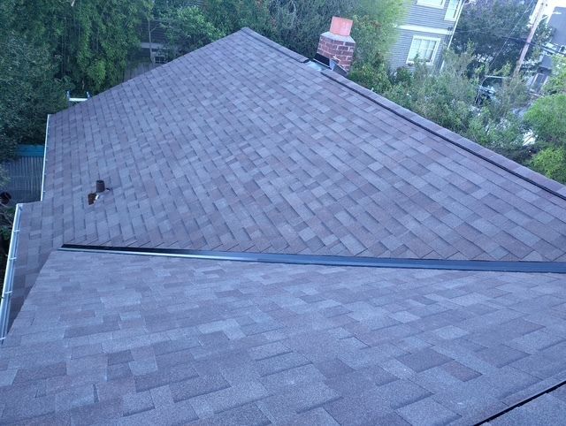 Gray shingled roof with two sections, viewed from above, surrounded by trees and houses