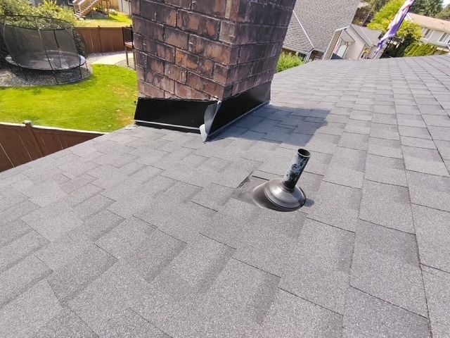 Rooftop shingles with a vent pipe near a brick chimney and a grassy yard in the background