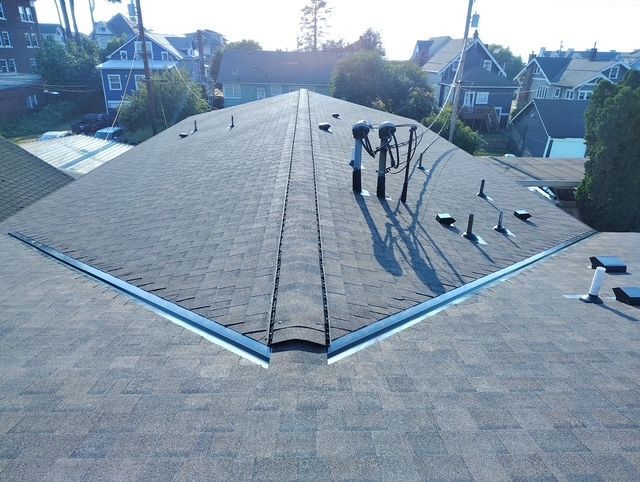 Roofers working on a gray shingled house roof, installing metal flashing near the ridge