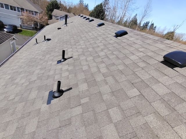 Tilted view of a shingled roof with vents and rooftop equipment near houses and trees