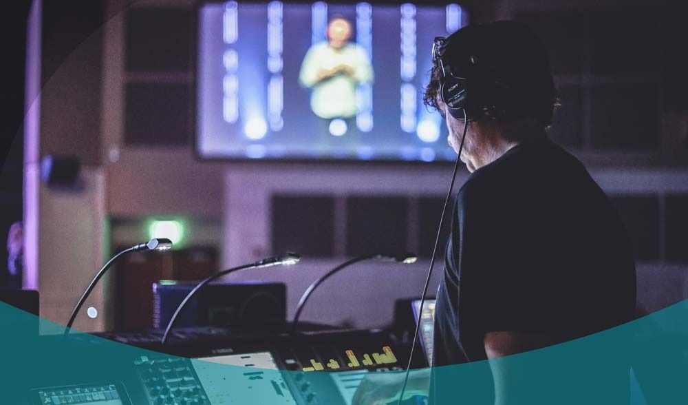 Una persona con le cuffie lavora a un mixer audio in uno studio scarsamente illuminato, con uno schermo da palcoscenico sullo sfondo.