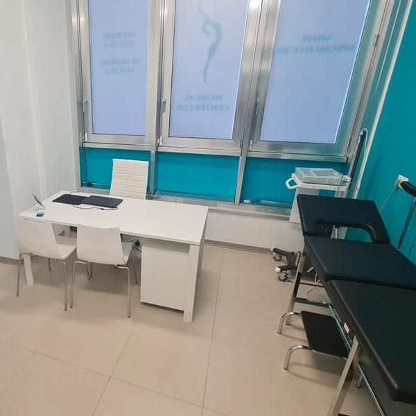 Uno studio medico con scrivania bianca, sedie e lettino da visita nero su un pavimento piastrellato chiaro.