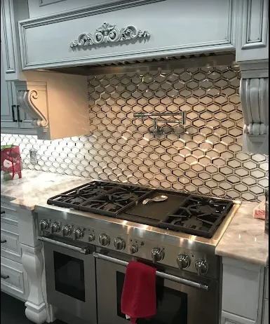 White kitchen with a stainless steel gas range, ornate hood, and patterned tile backsplash.