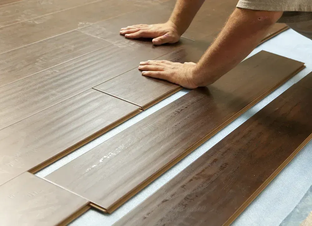 Hands installing dark wood floor planks beside a silver underlayment sheet.