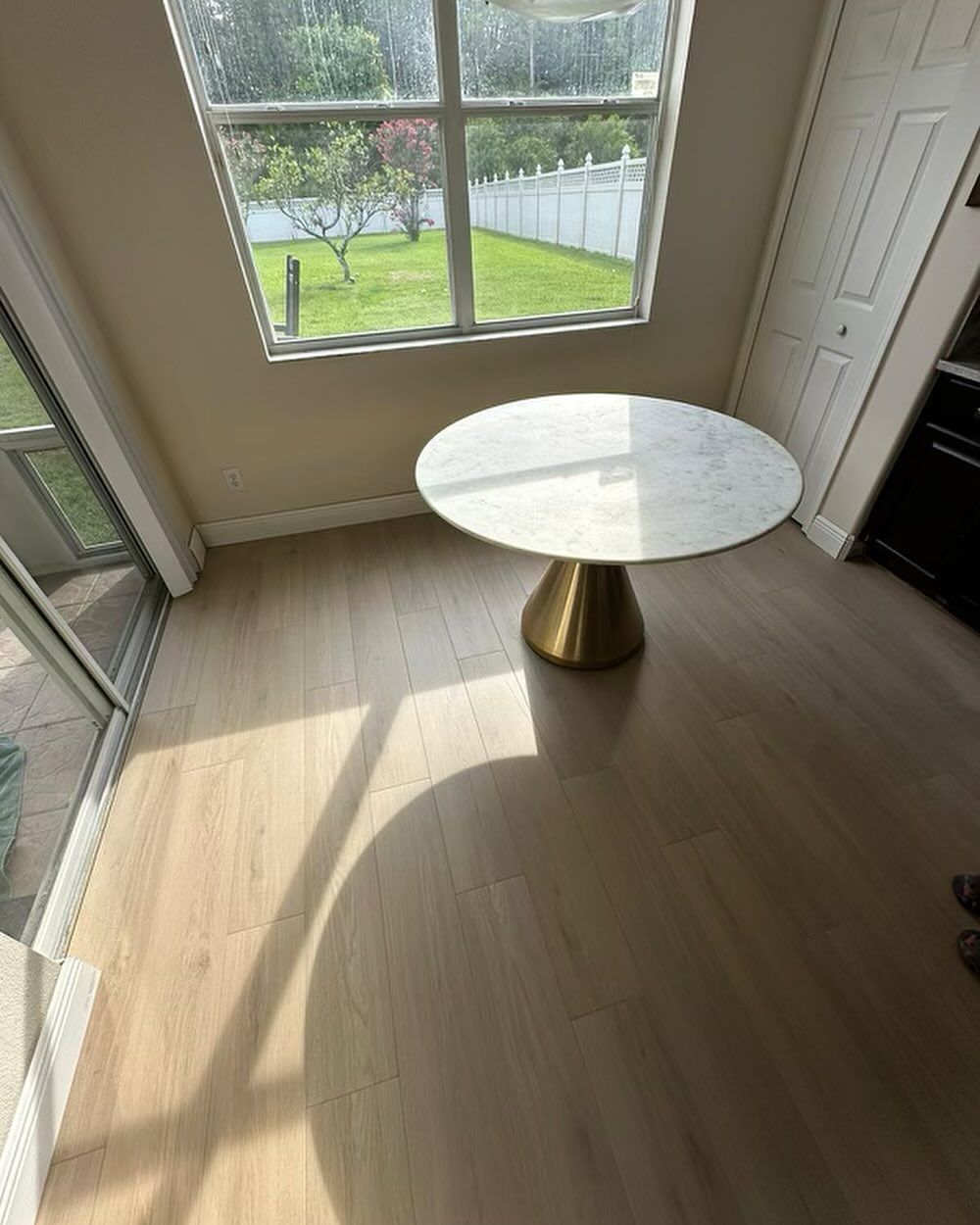 Sunlit empty room with large windows, light wood floor, and a round white table on a gold pedestal base