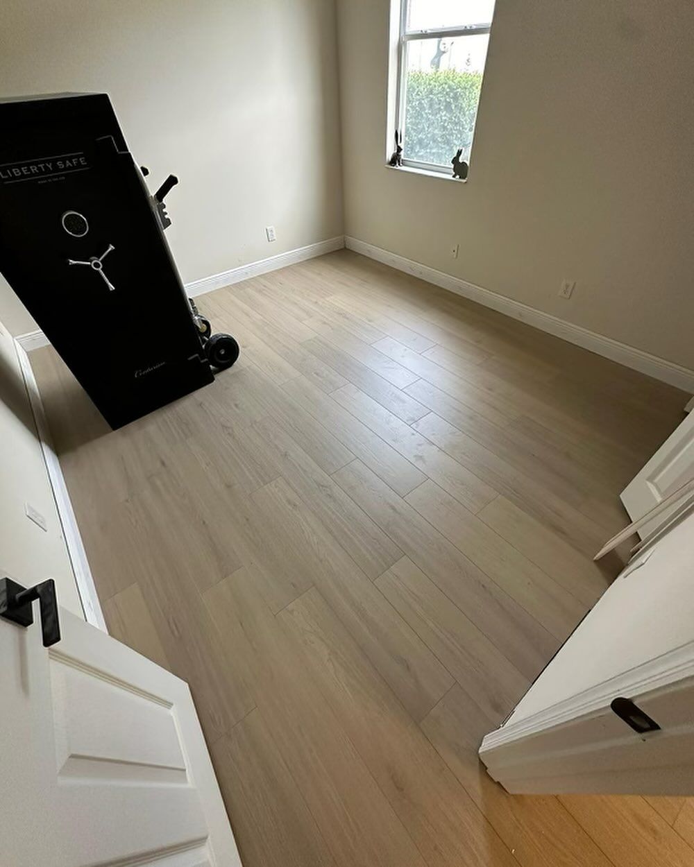 Empty beige bedroom with light wood floor, open white doors, and a black dresser by the window.