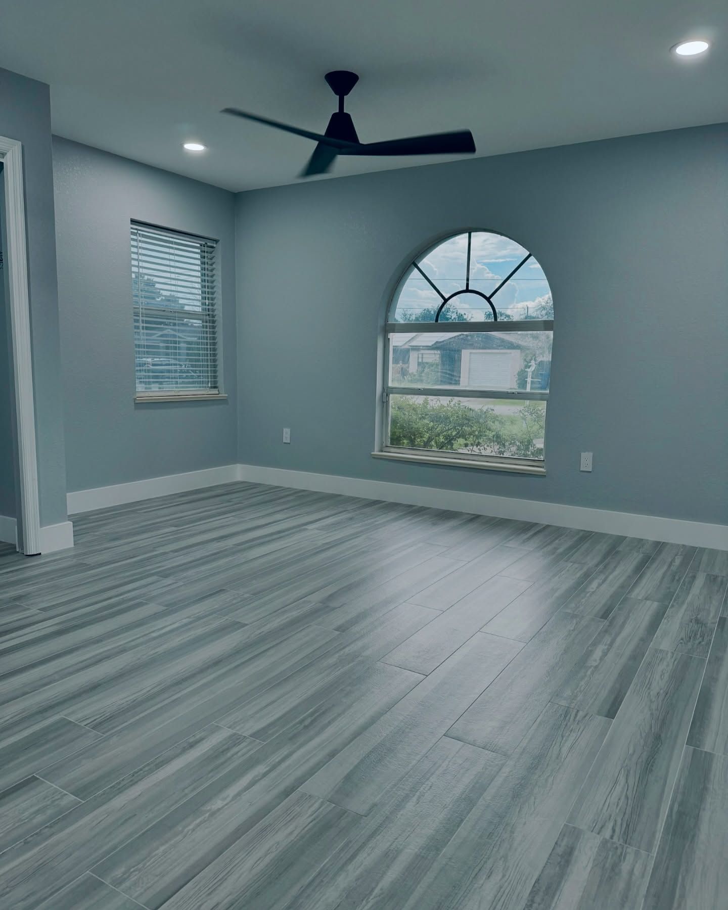 Empty blue-gray room with wood-look floors, arched window, ceiling fan, and white trim