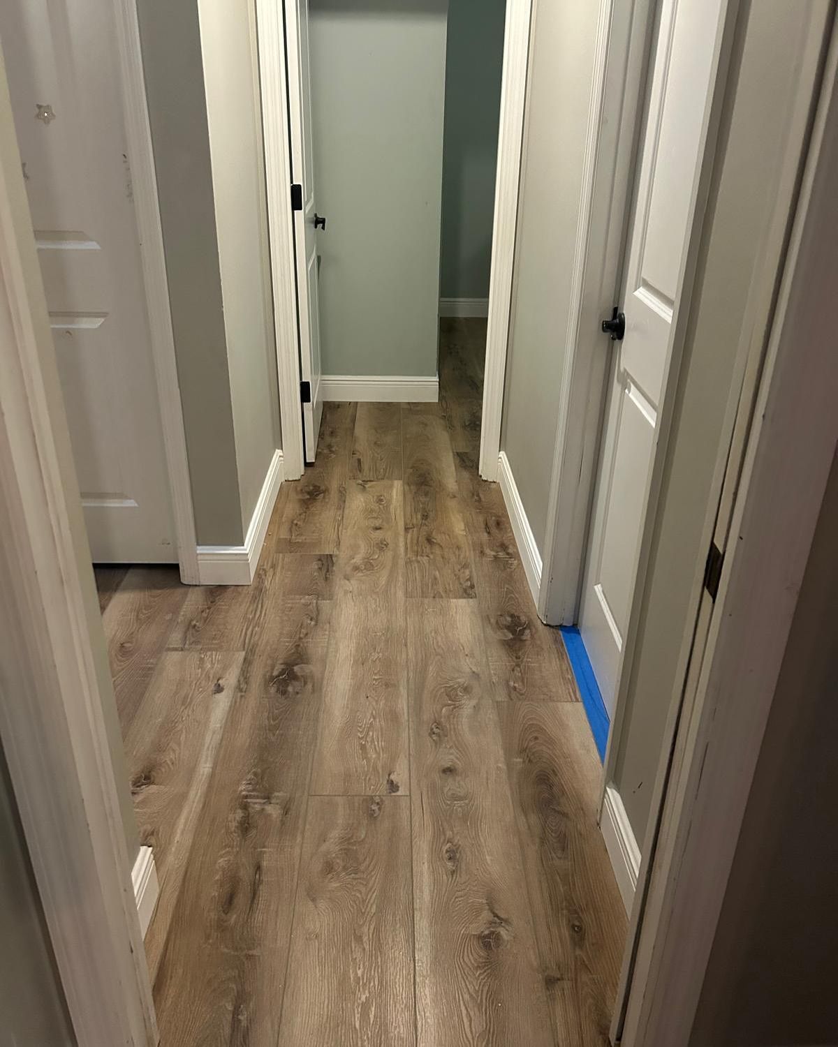 Narrow hallway with wood-look flooring, white doors, and blue painter’s tape near the baseboards